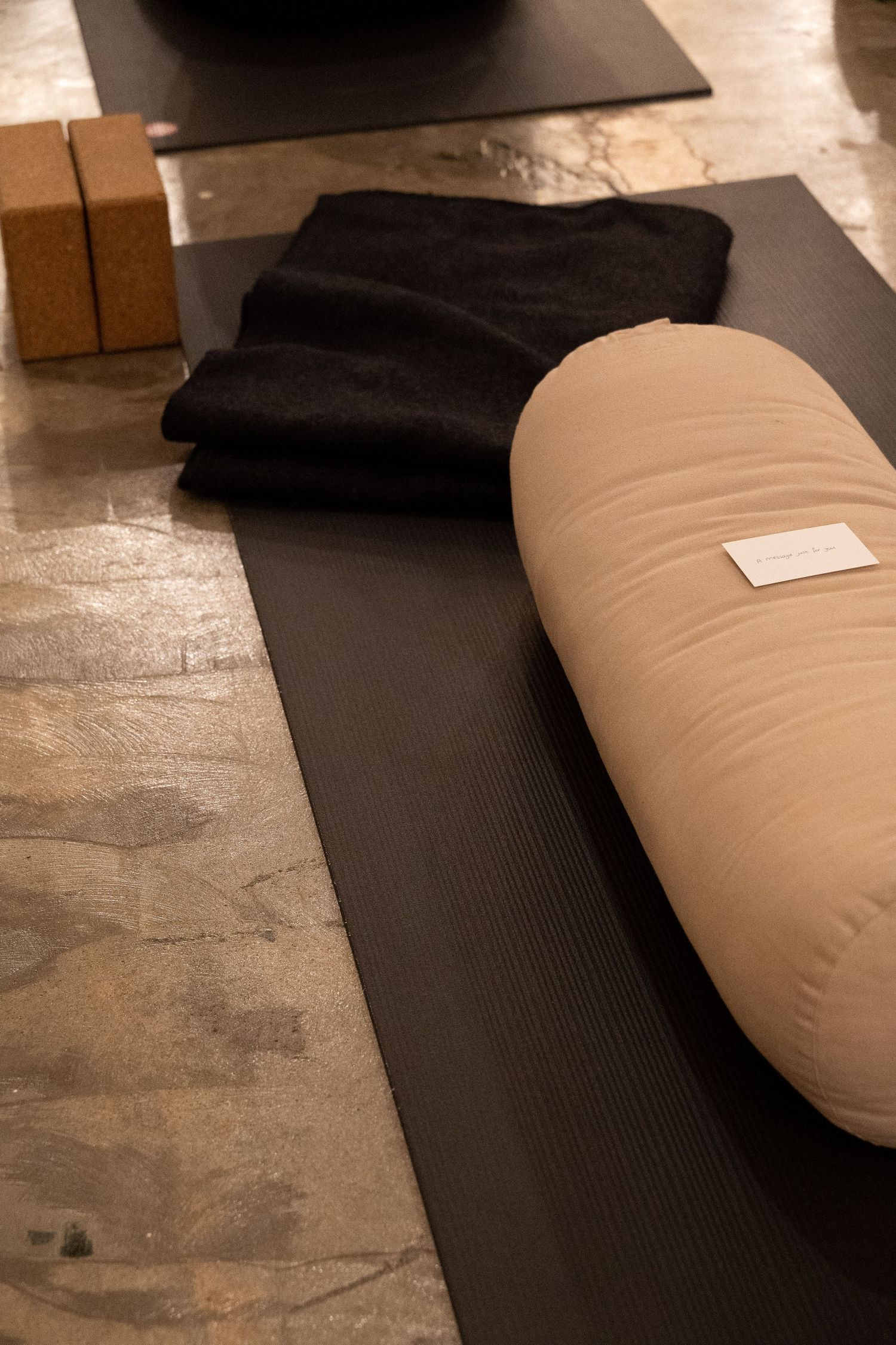 Yoga mat with beige bolster pillow, black blanket, and cork blocks on studio floor.