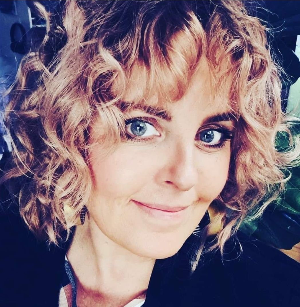 Woman with short curly blonde hair and blue eyes smiling in a close-up selfie portrait.