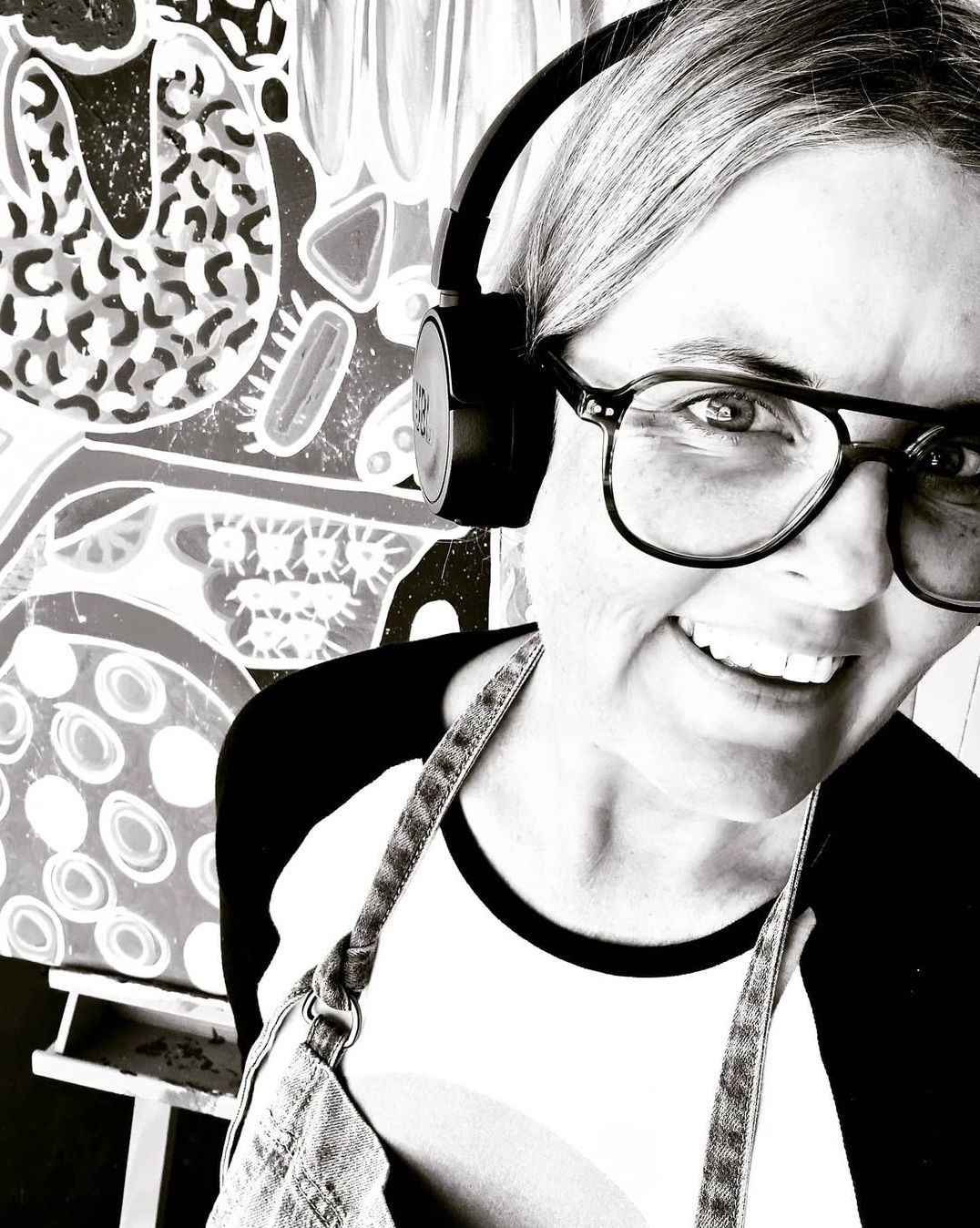 Black and white selfie of a smiling woman wearing headphones and glasses in an art studio.