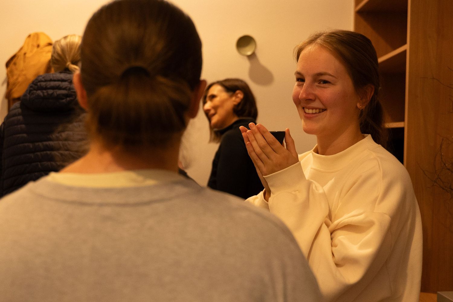 Two women smiling and greeting each other warmly inside a cozy yoga studio changing room.