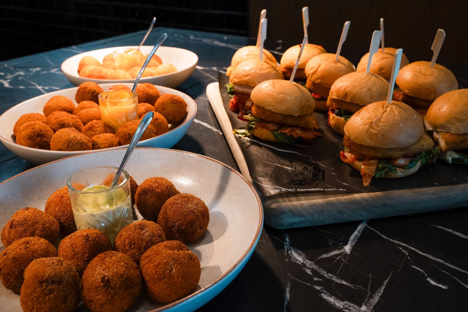 Assorted appetizers including fried croquettes, mini sliders on a wooden board, and a bowl of fruit on a marble table.