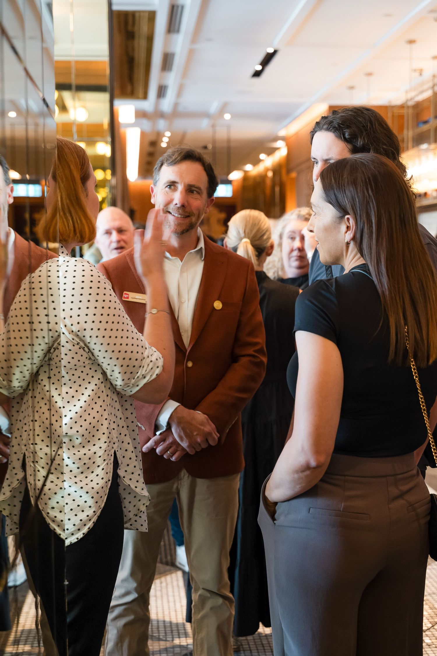 Professionals networking at an indoor event, man in brown blazer conversing with women in modern venue.