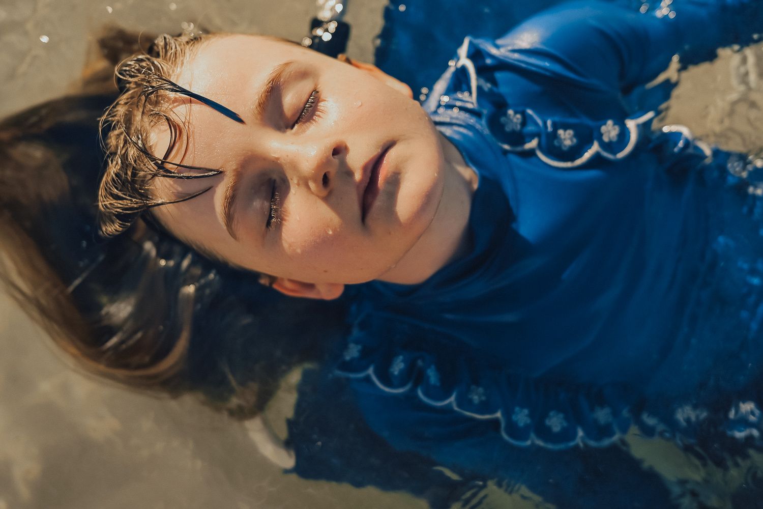 Woman in blue costume lying in water with dramatic eye makeup and wet hair, artistic fashion photography.