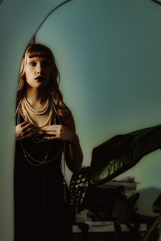Woman in black dress with pearl necklaces and headband poses elegantly in moody green-lit studio setting.