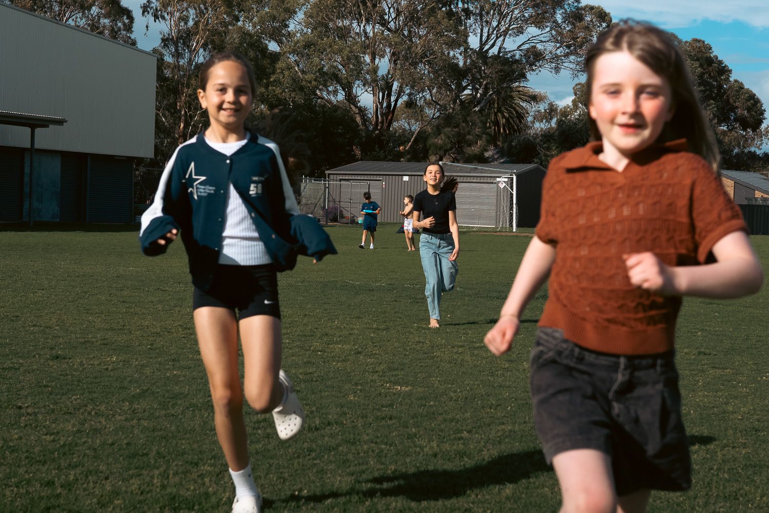 Girls running and laughing on a sunny green school oval during outdoor playtime.