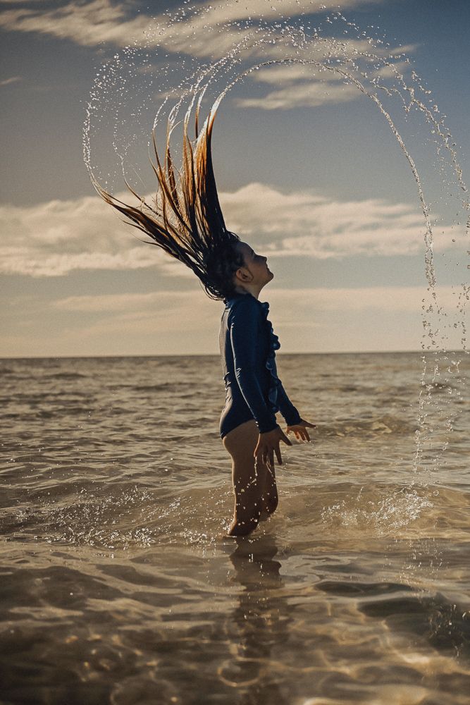 Girl in blue swimsuit flipping wet hair creating water arc while standing in ocean waves at sunset.