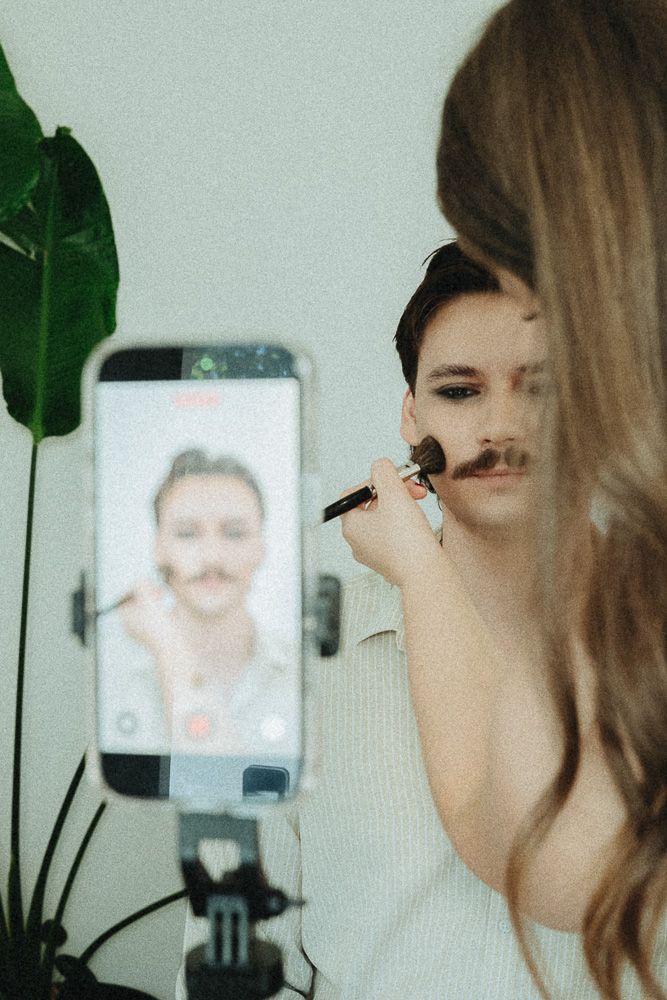 Woman applying makeup with brush on person with mustache, filmed live on smartphone mounted on tripod.