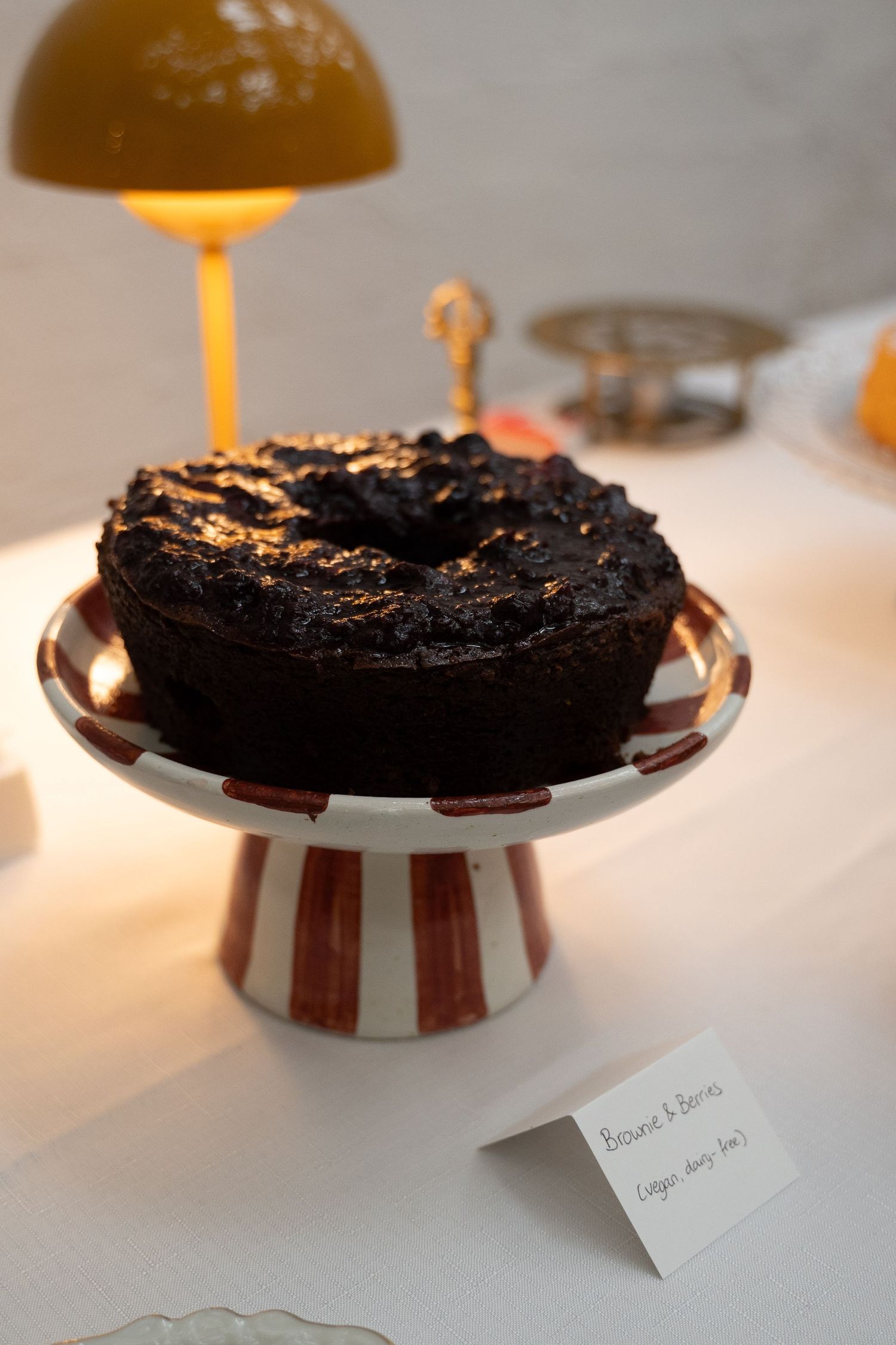 Dark chocolate bundt cake on striped red and white cake stand, illuminated by warm amber lamp on white table.