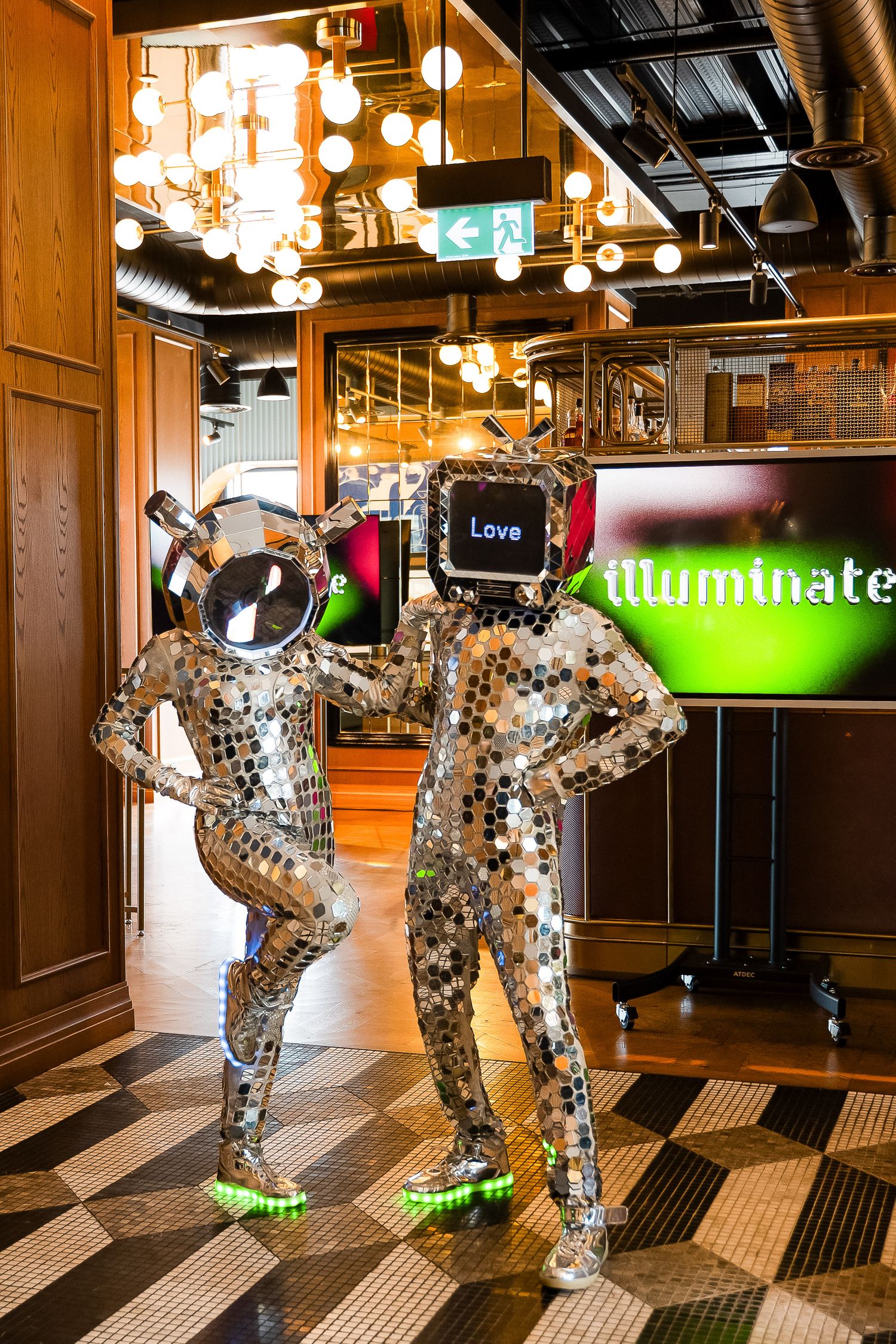 Two performers in mirrored disco ball suits pose indoors near an Illuminate sign, one wearing a helmet, the other a TV head displaying Love.