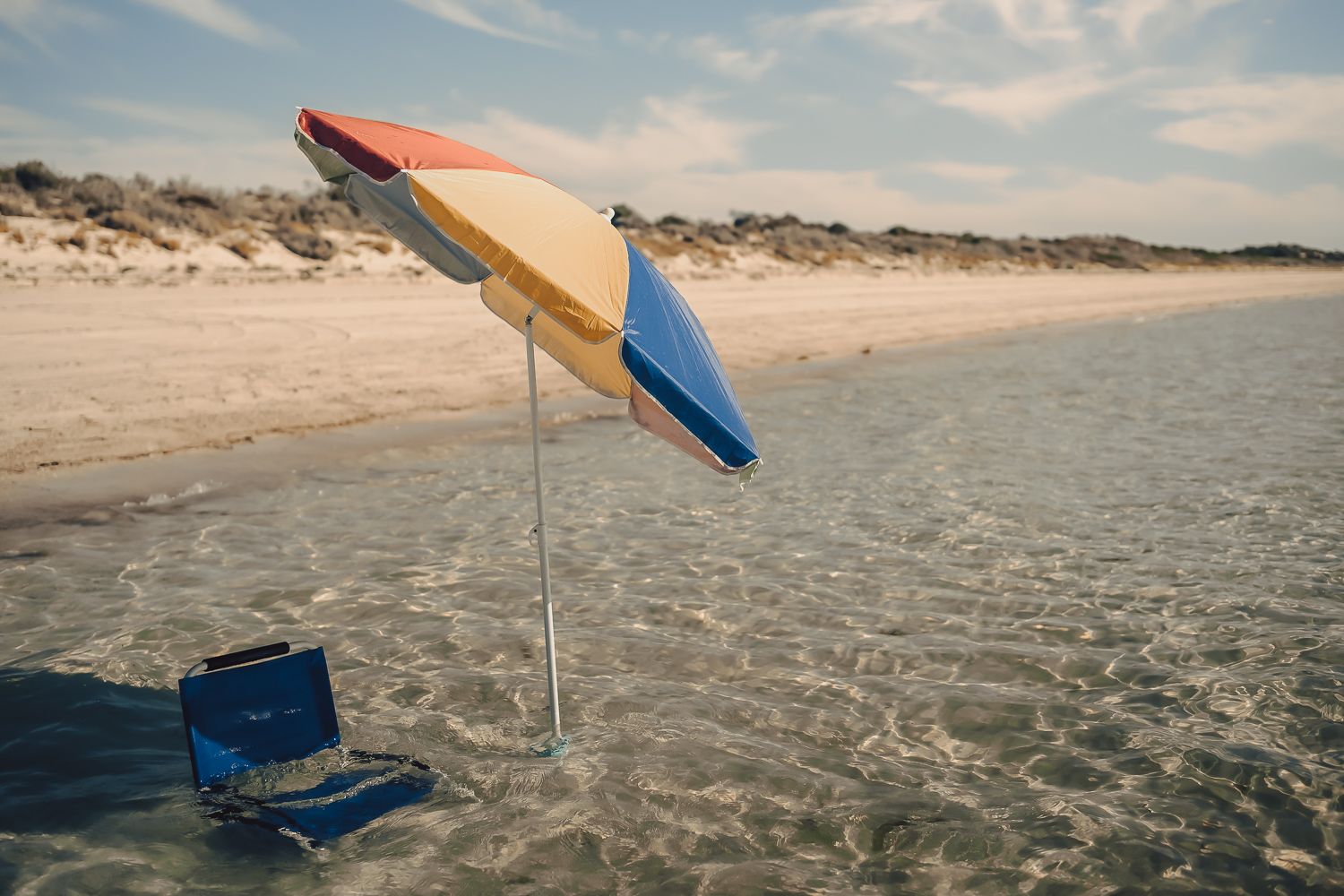 Colorful beach umbrella standing in shallow ocean water beside a blue bag on a sandy shore.