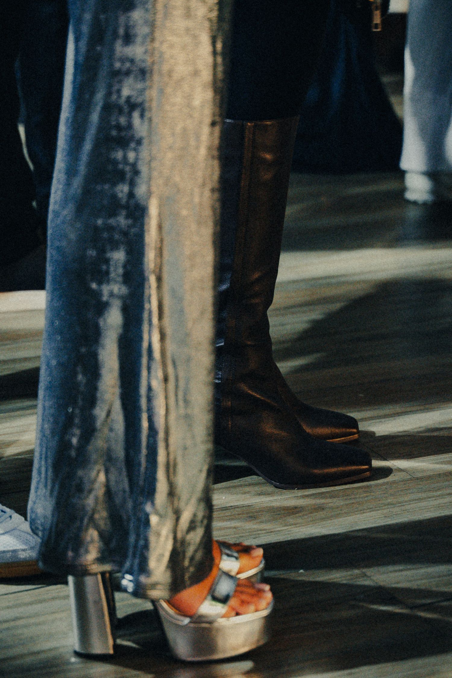 Close-up of fashion footwear featuring a tall black leather boot and orange platform sandal on wooden floor.