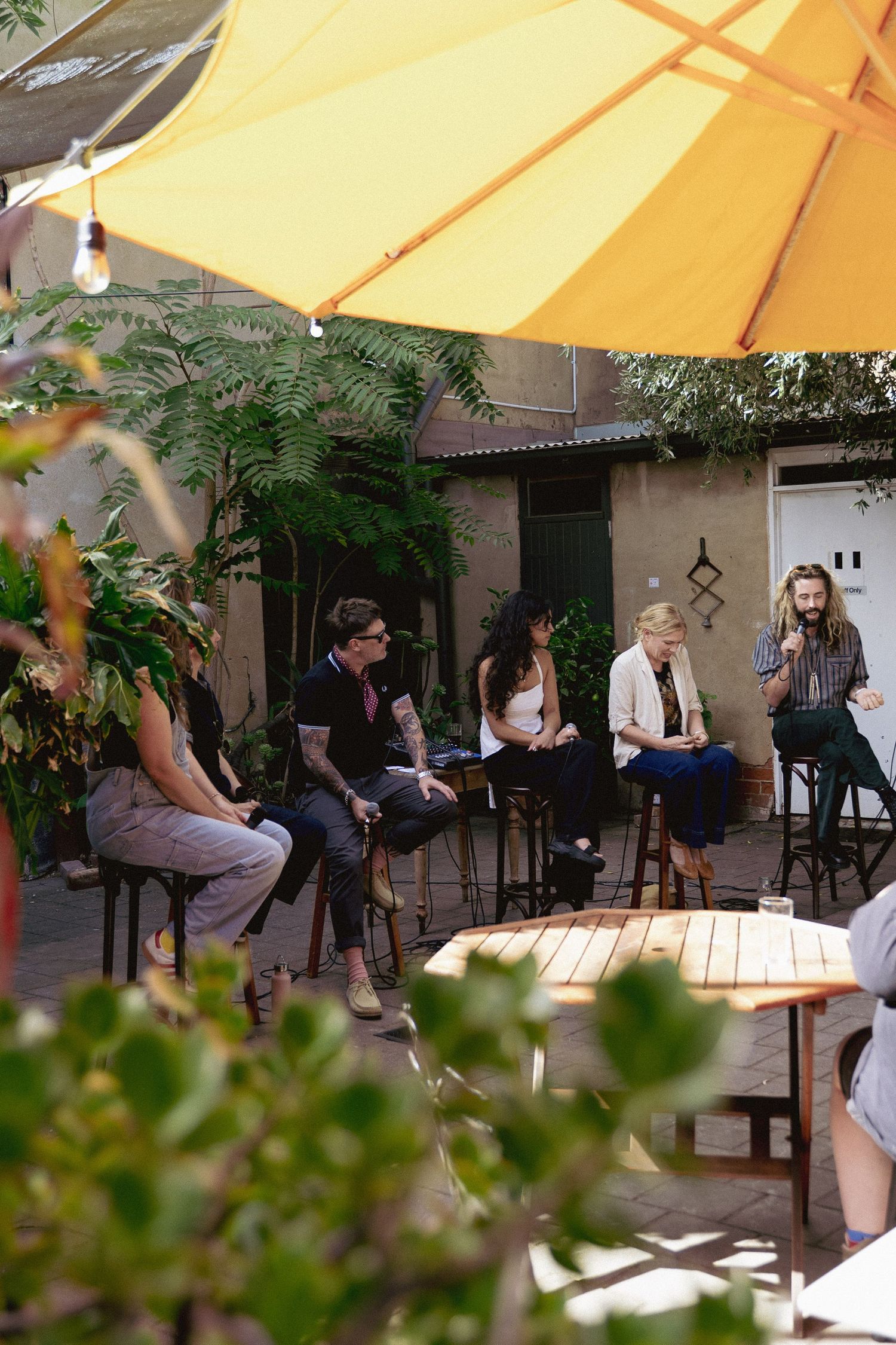 Group of friends enjoying outdoor cafe conversation under yellow umbrella surrounded by lush greenery.