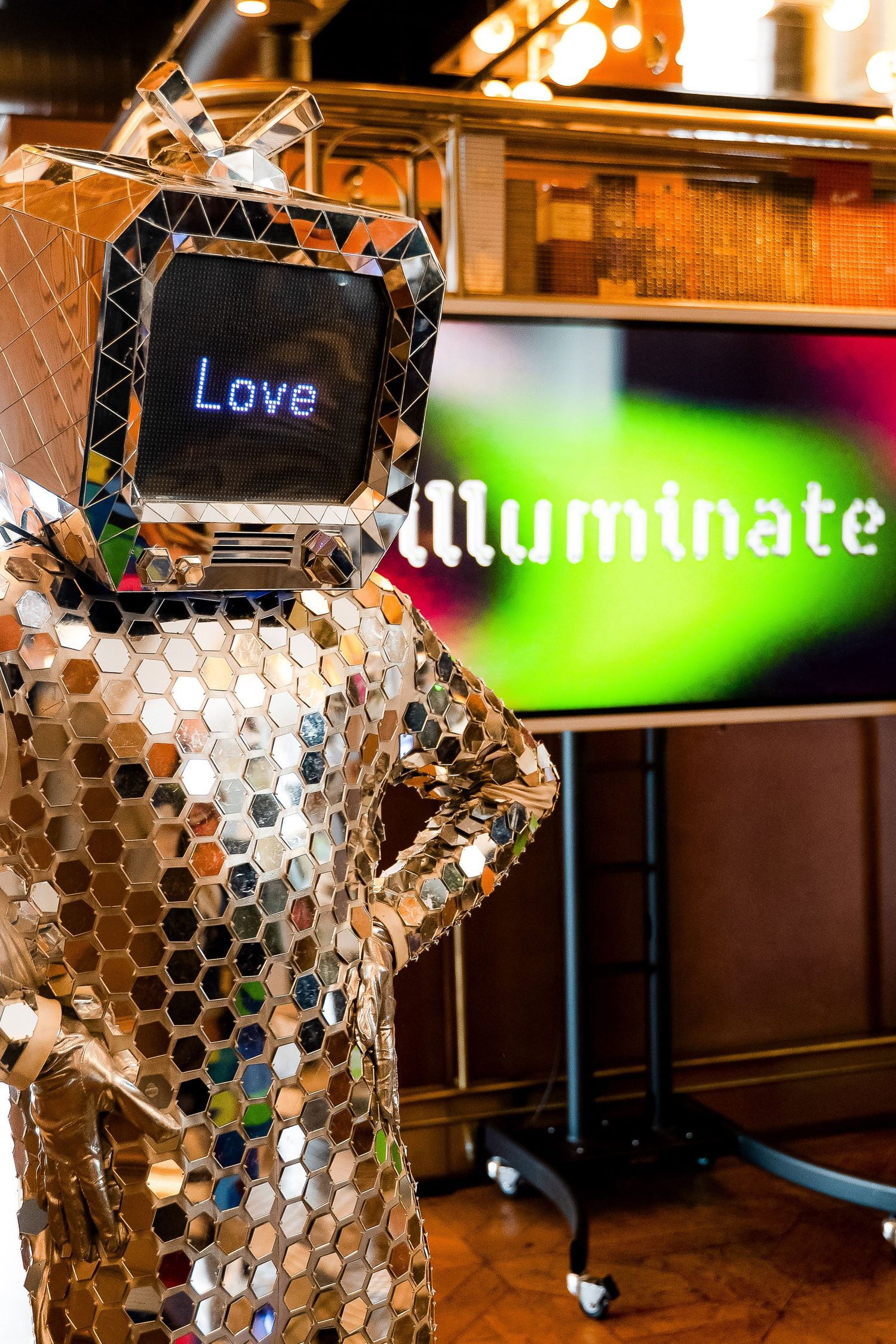 A mirror-covered robot costume with a TV head displaying 'Love' stands before an Illuminate event screen.