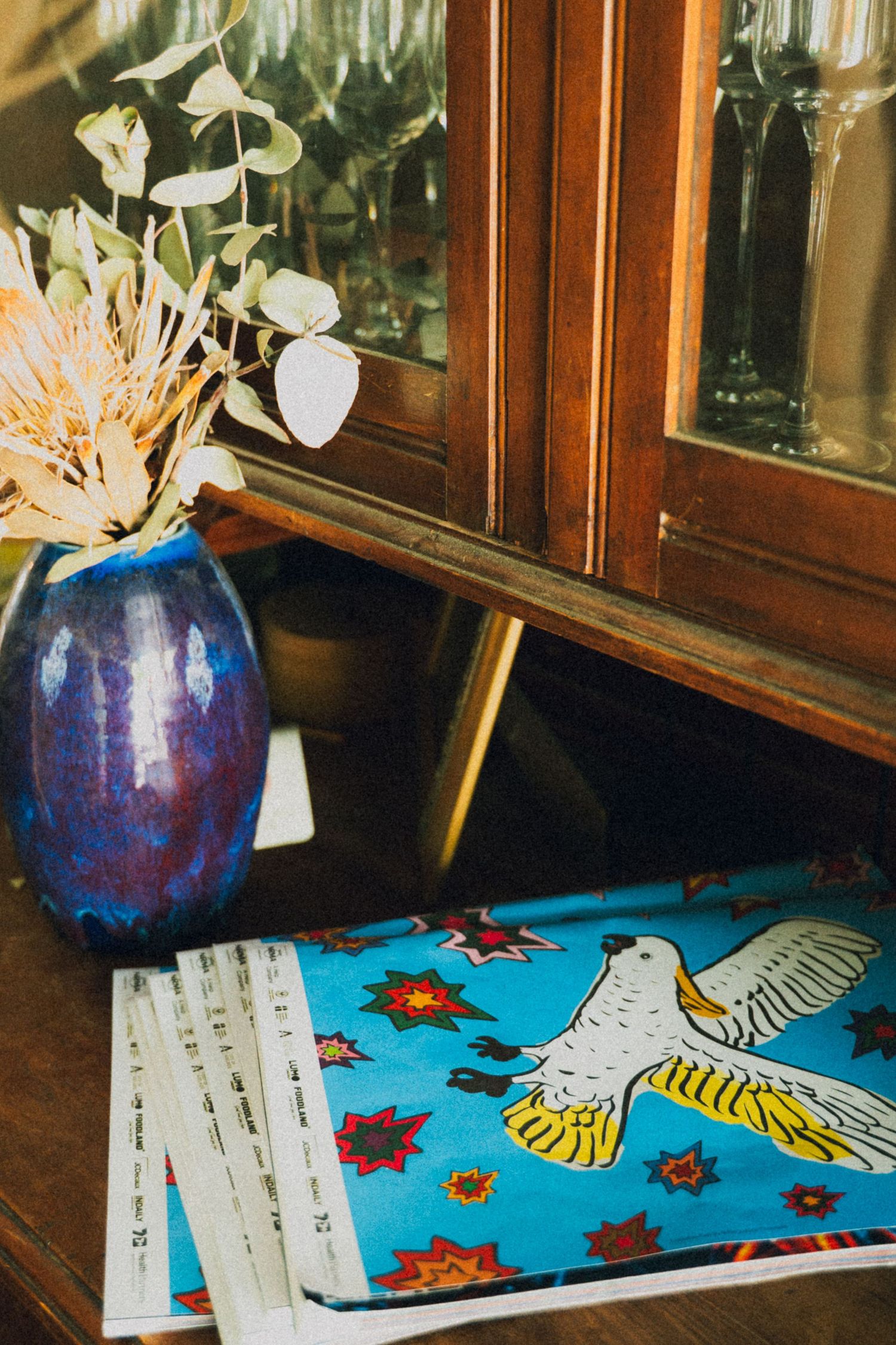 Blue ceramic vase with dried flowers beside a colorful bird-printed book on a wooden sideboard.