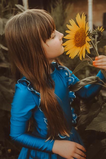 Young girl in blue dress smelling a bright yellow sunflower in a garden setting.