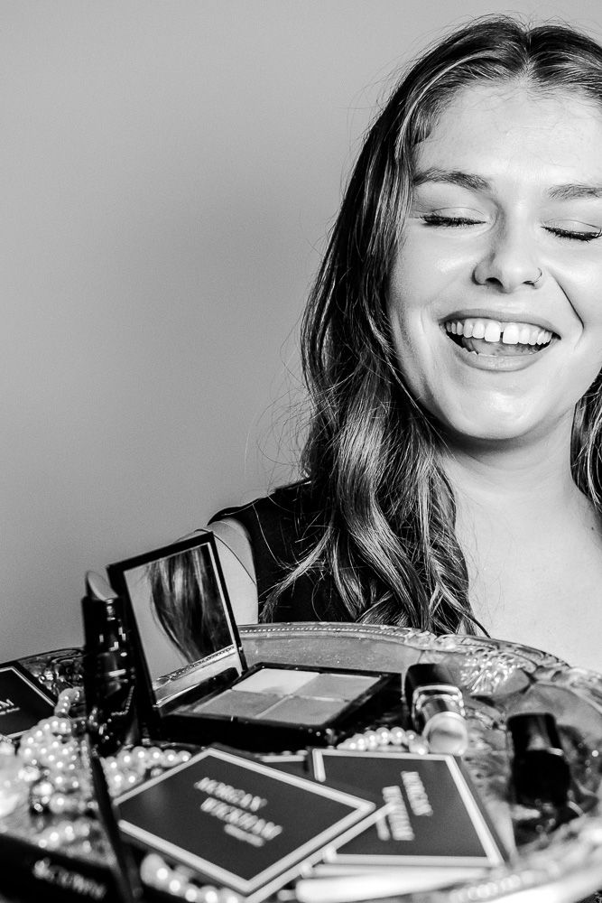 Black and white photo of a smiling young woman with makeup products and jewelry displayed in front of her.