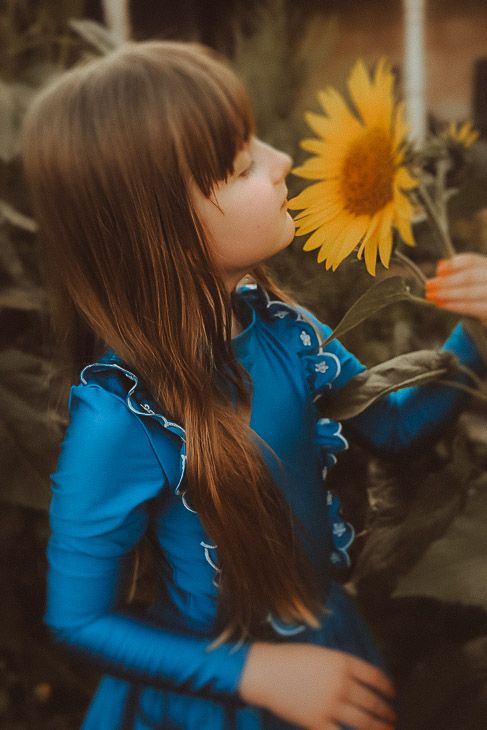 A young girl in a blue dress with long brown hair smelling a bright yellow sunflower outdoors.