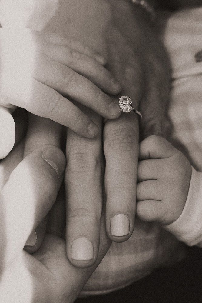 Black and white photo of adult hands and a baby's hand, with a diamond engagement ring on the woman's finger.
