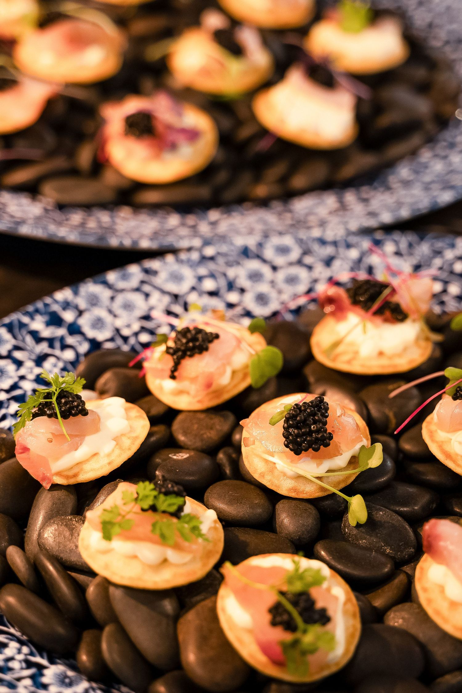 Elegant canapes with caviar, smoked salmon, and herbs served on black river stones at a catered event.