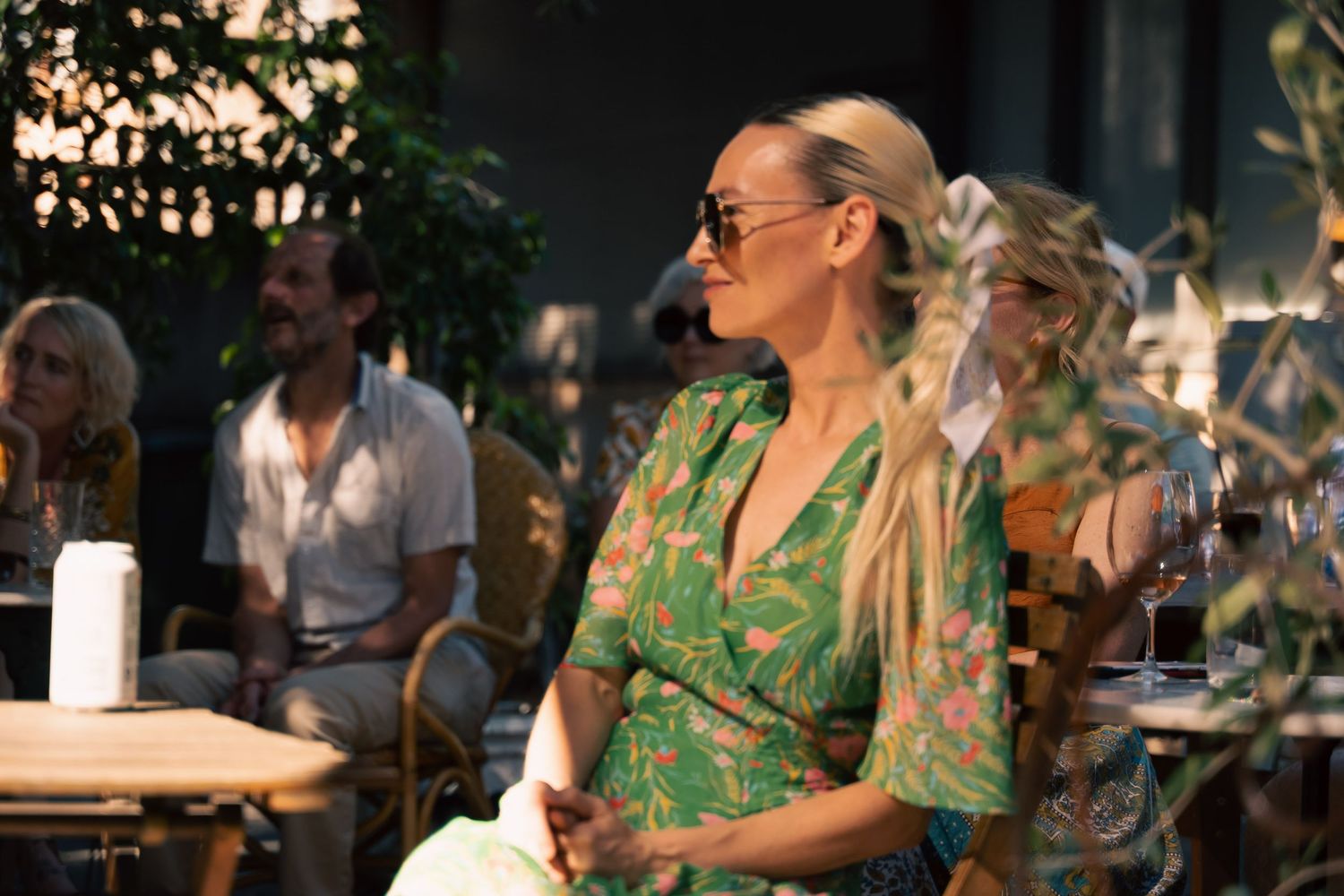 Blonde woman in green floral dress and sunglasses sitting outdoors at a cafe patio in sunlight.