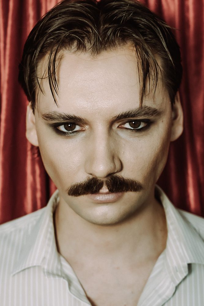 Portrait of a man with a mustache, dark eye makeup, slicked hair, and a white shirt against red curtains.
