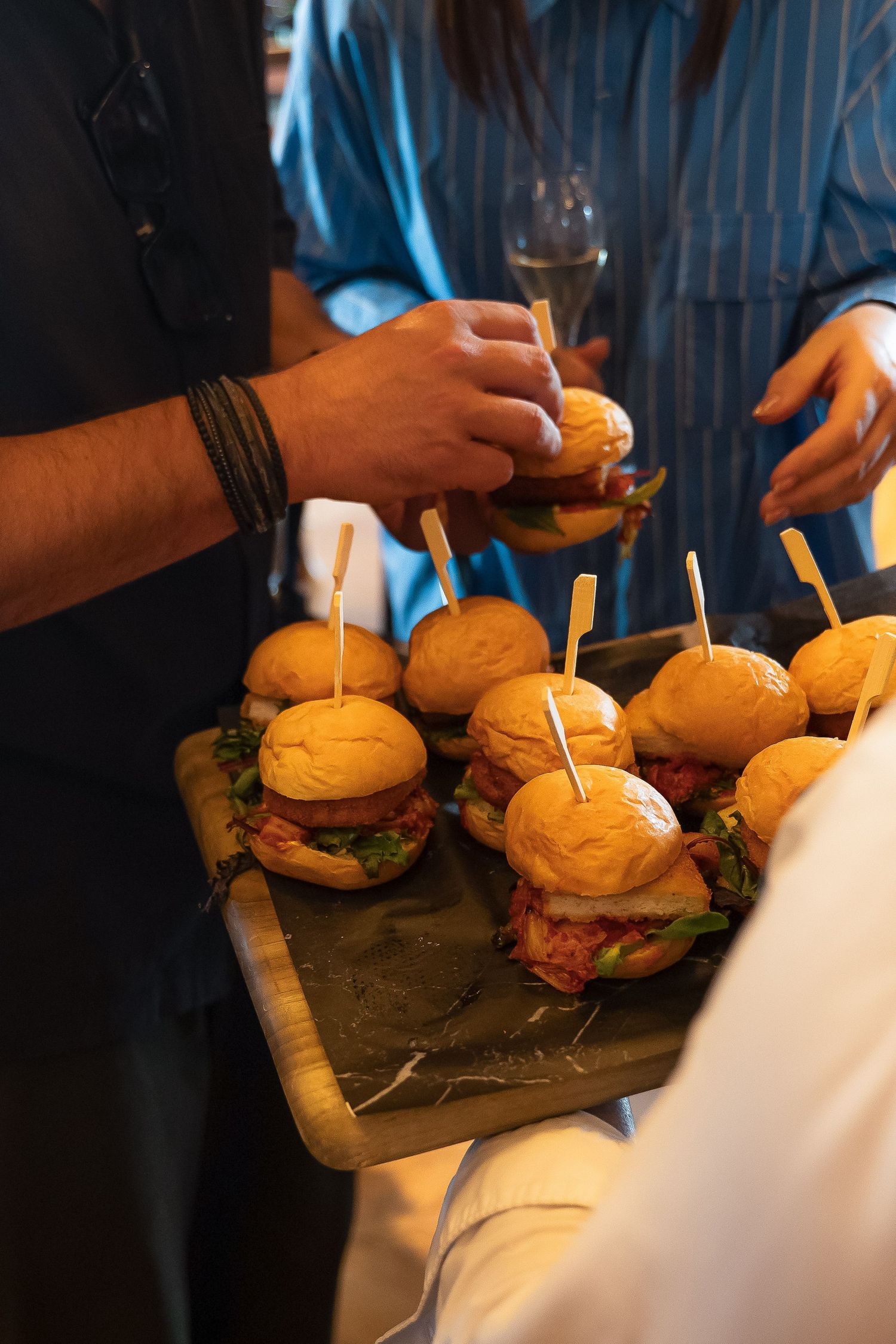 A server presents a wooden board of mini sliders with toothpicks at a catered event while guests reach for the appetizers.