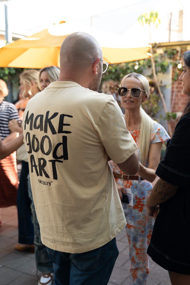 Man wearing a cream 'Make Good Art' t-shirt at an outdoor social gathering with people and yellow umbrellas.