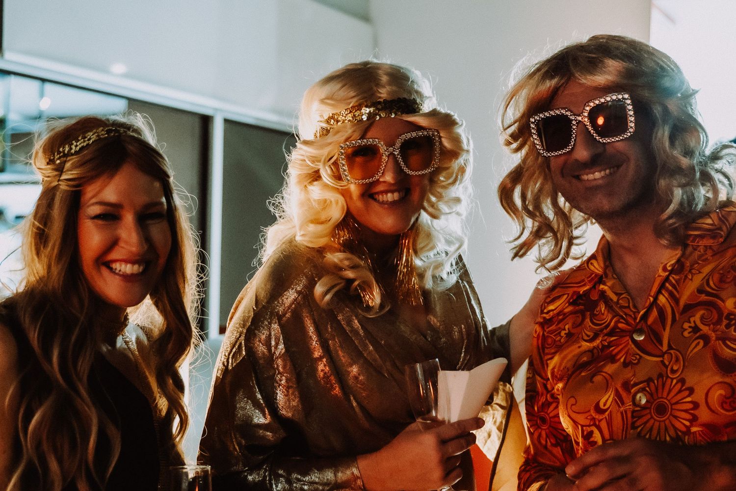 Three women in 1970s retro costumes with oversized sunglasses and wigs smile together at a themed party.