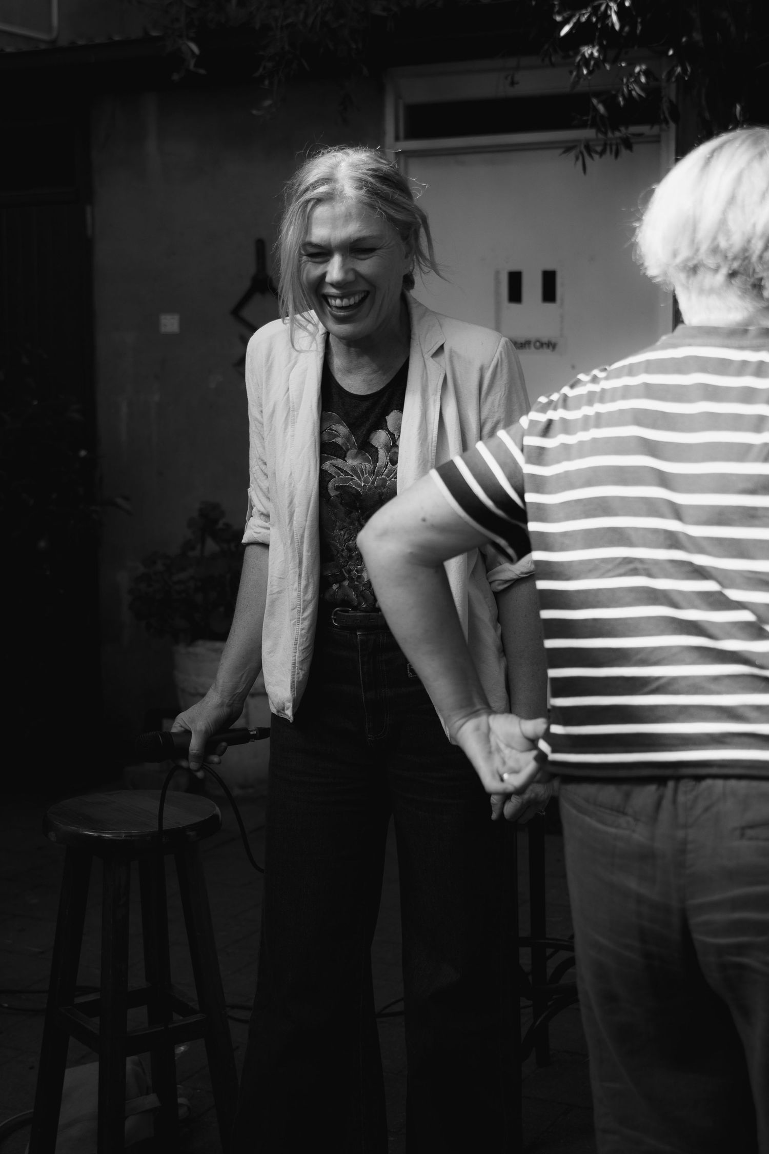 Black and white photo of a laughing blonde woman in a blazer holding a stool, shaking hands with a person in a striped shirt.