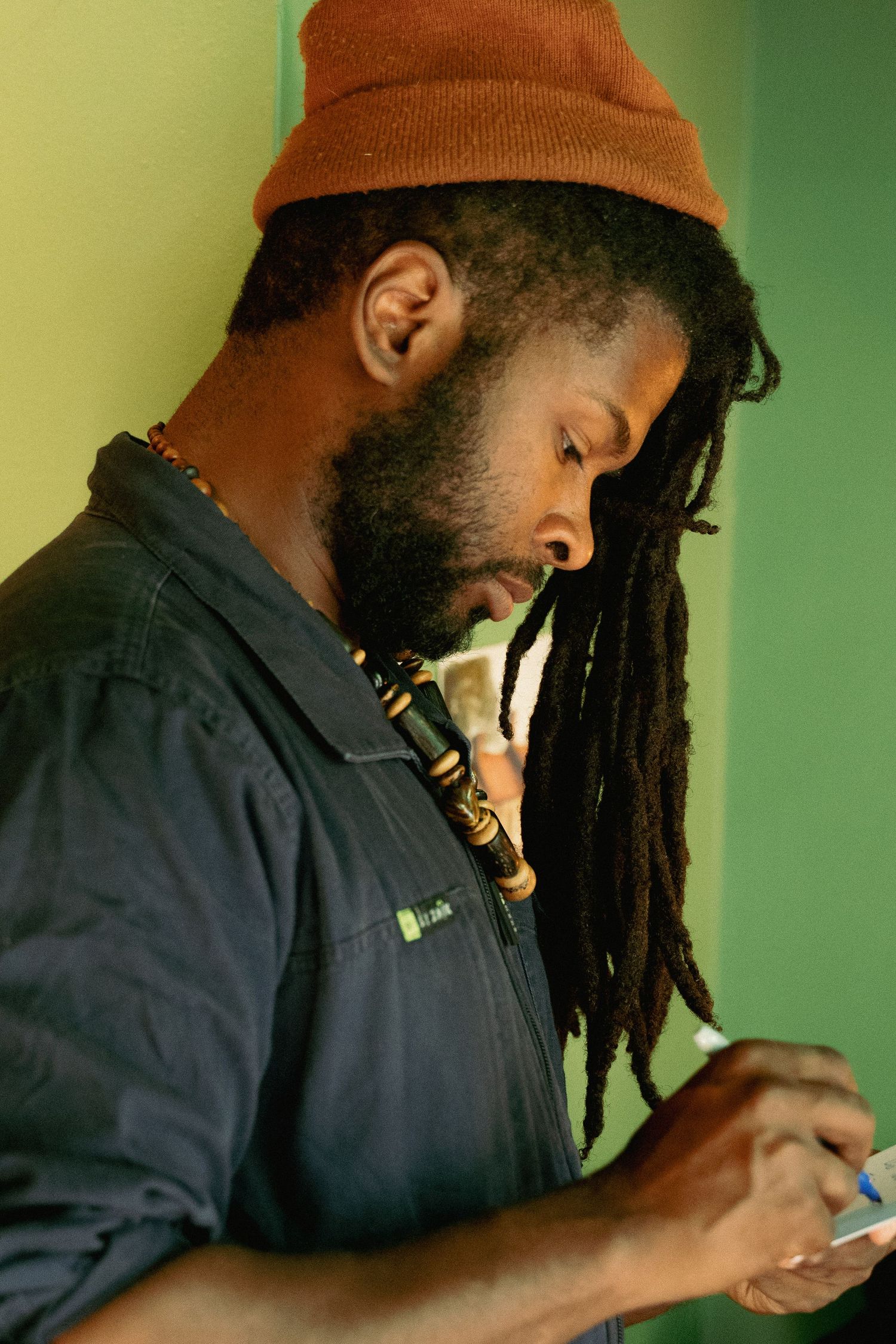 Man with dreadlocks and orange beanie writing notes while leaning against a green wall.