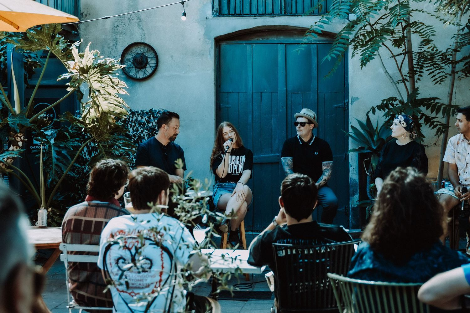 Group of people gathered in a courtyard for an outdoor discussion panel, surrounded by lush plants and a blue door.
