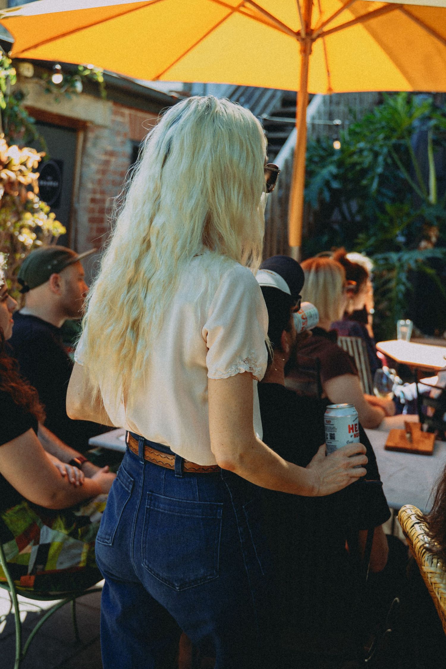 Blonde woman in white top and jeans holds a can at an outdoor patio gathering under a yellow umbrella.