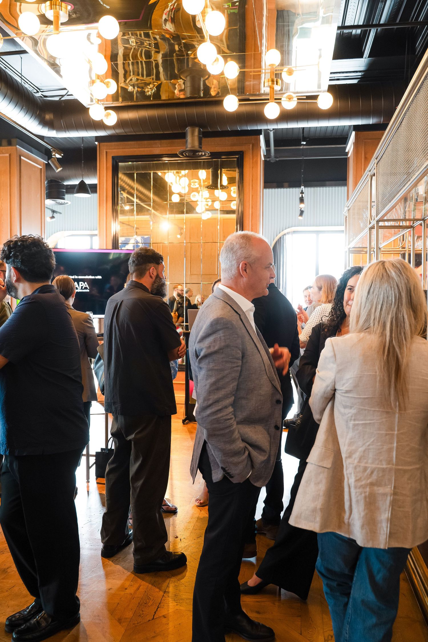 Professionals networking at an upscale indoor event venue with warm Edison bulb lighting and wooden floors.