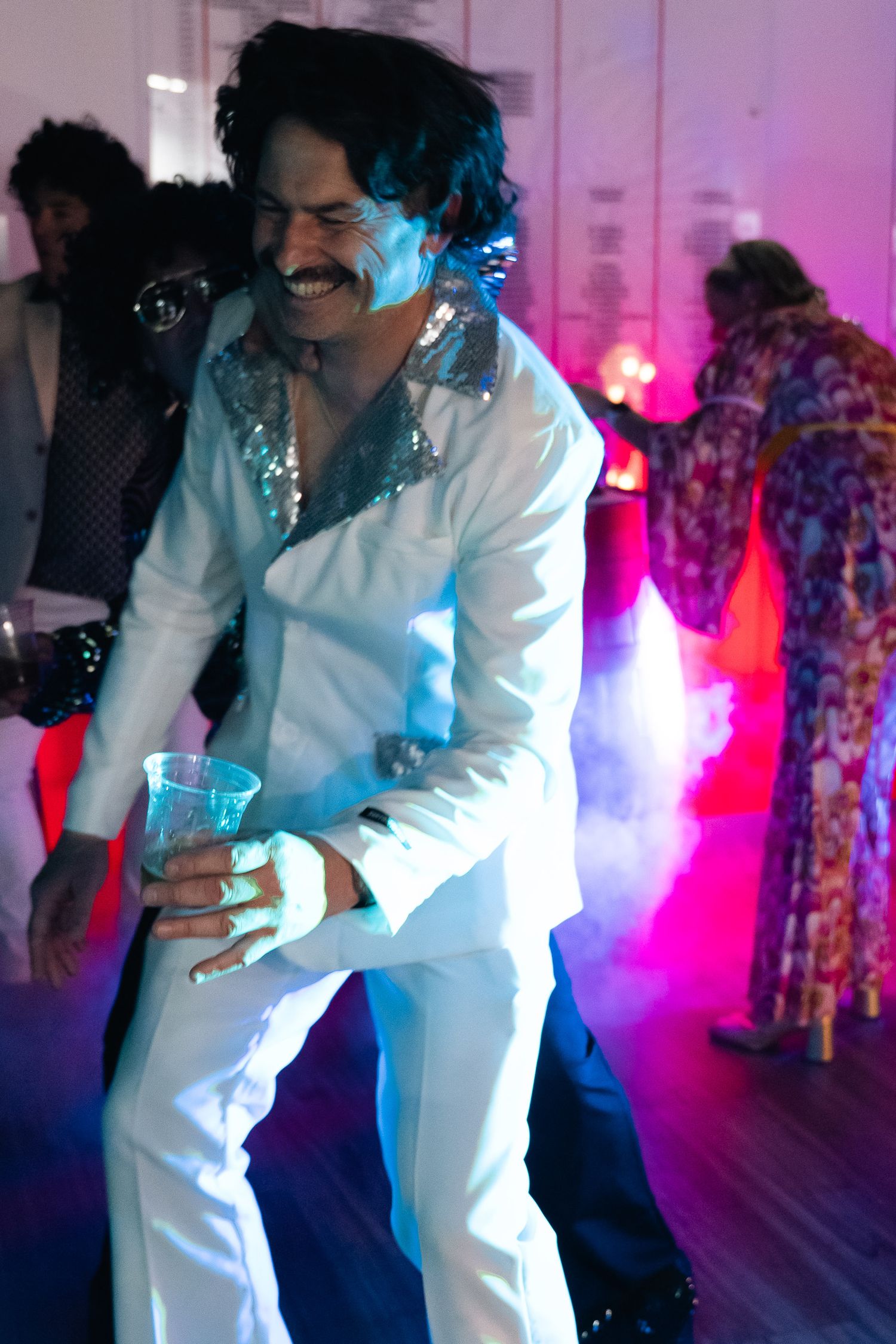 Man in white disco suit with sequin collar dancing at a retro 70s themed party with colorful lights.