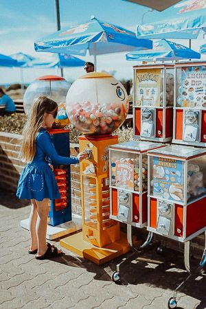 Young girl in blue dress using colorful gumball vending machines at outdoor amusement area.
