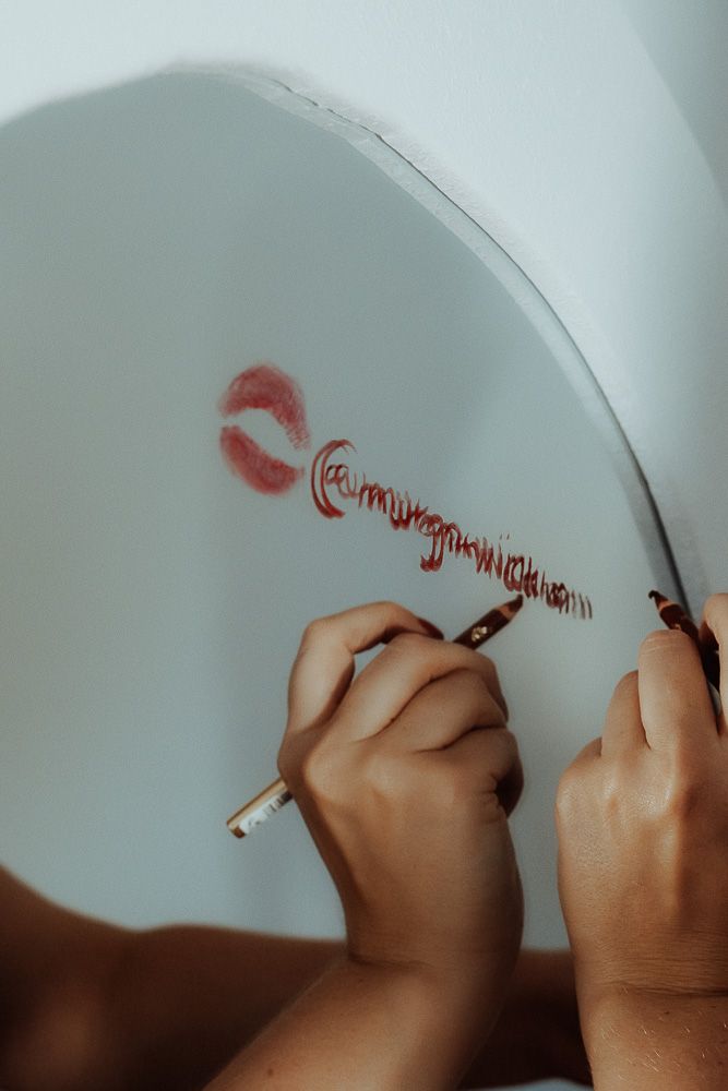 Hands writing in red lipstick on a mirror with a lip print kiss mark beside the cursive text.