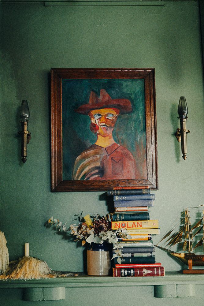 Vintage portrait painting in wooden frame above stacked books and ship model on green shelf with wall sconces.
