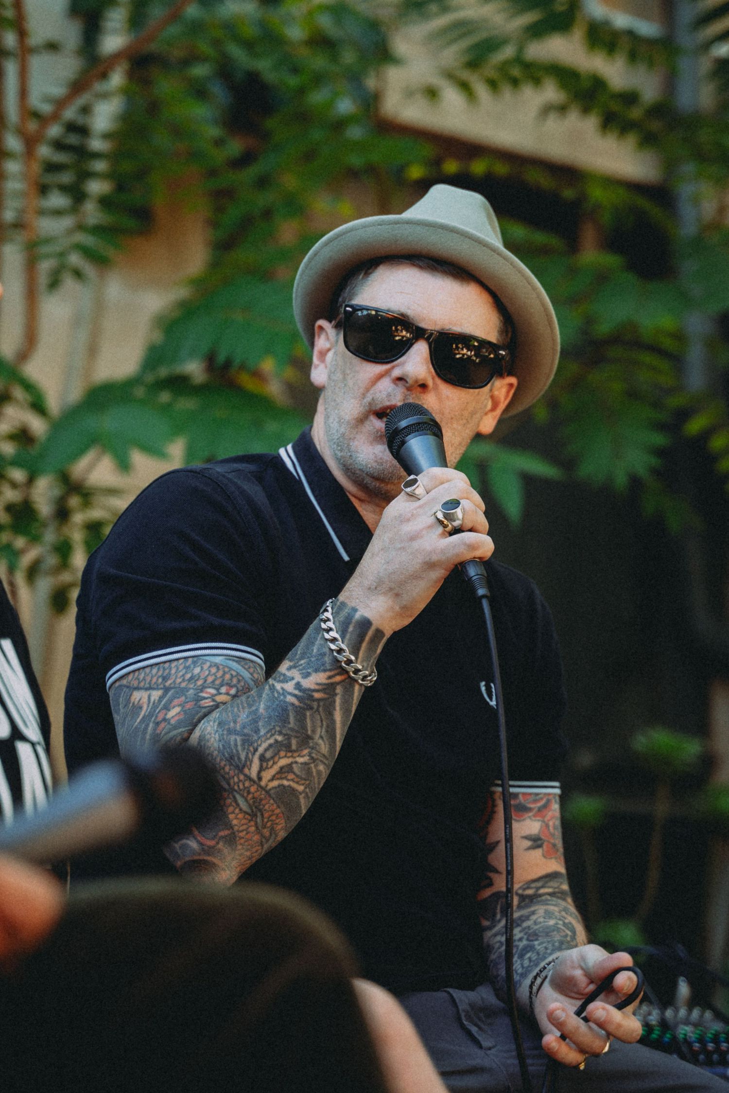 Tattooed man singing into microphone wearing black polo, gray fedora, and sunglasses performing outdoors.