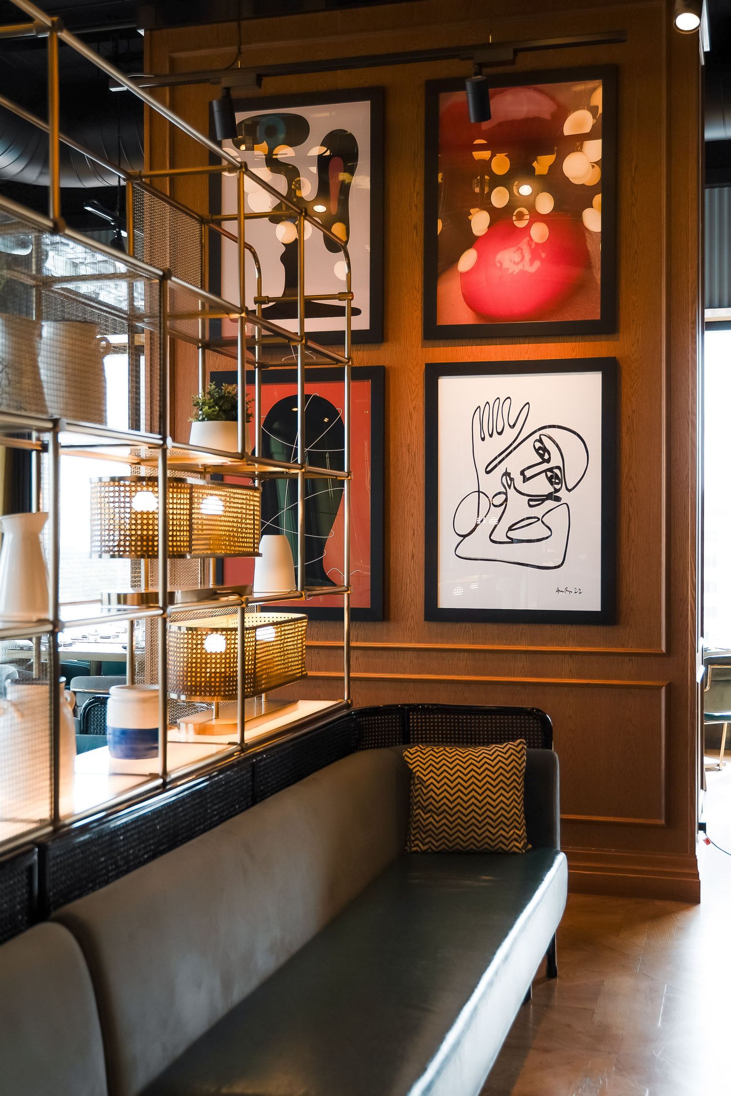 Modern lounge with gray velvet sofa, gold shelving unit with lamps, and eclectic framed artwork on warm wood-paneled walls.