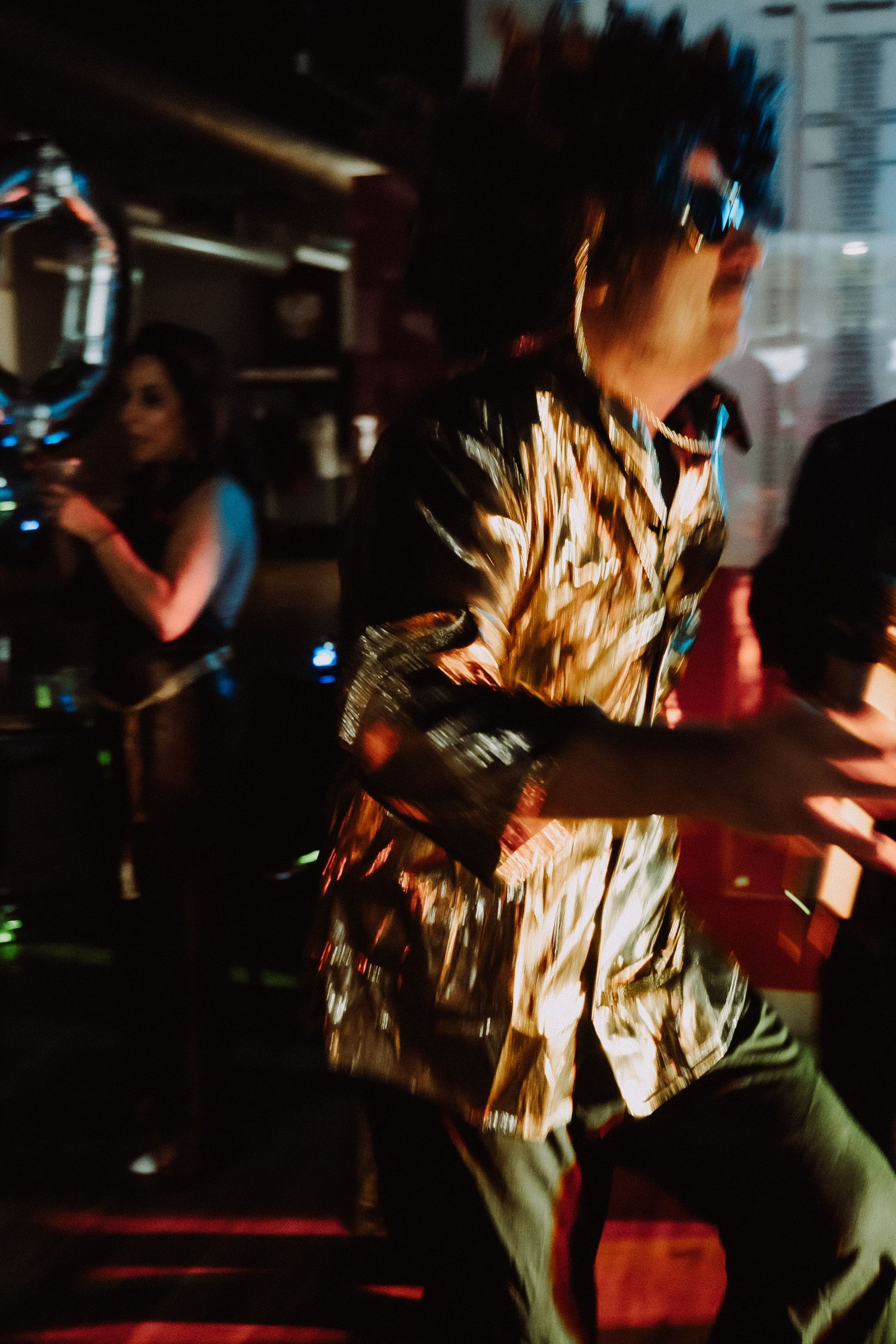 Blurred motion shot of a person dancing in a shiny gold shirt at a dark nightclub party.
