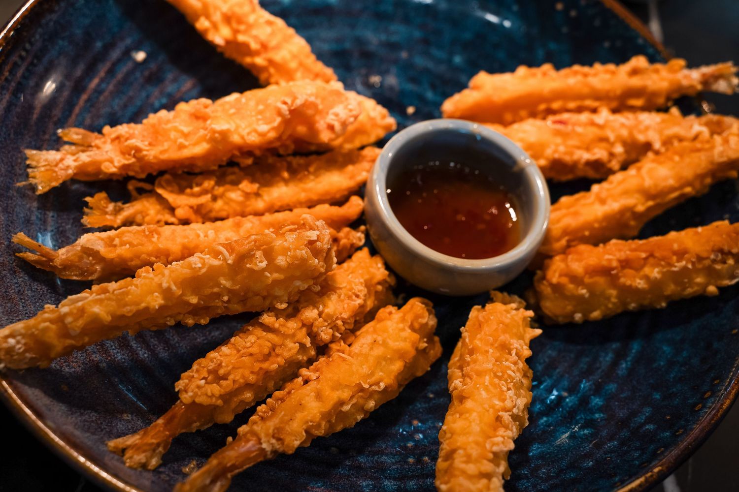 Golden crispy fried shrimp arranged on a blue plate with a small bowl of sweet chili dipping sauce.