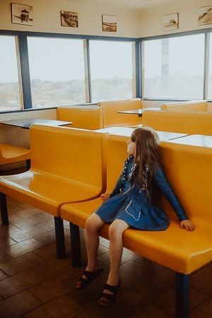 Young girl in blue dress sitting alone on yellow bench in a waiting room with large windows.