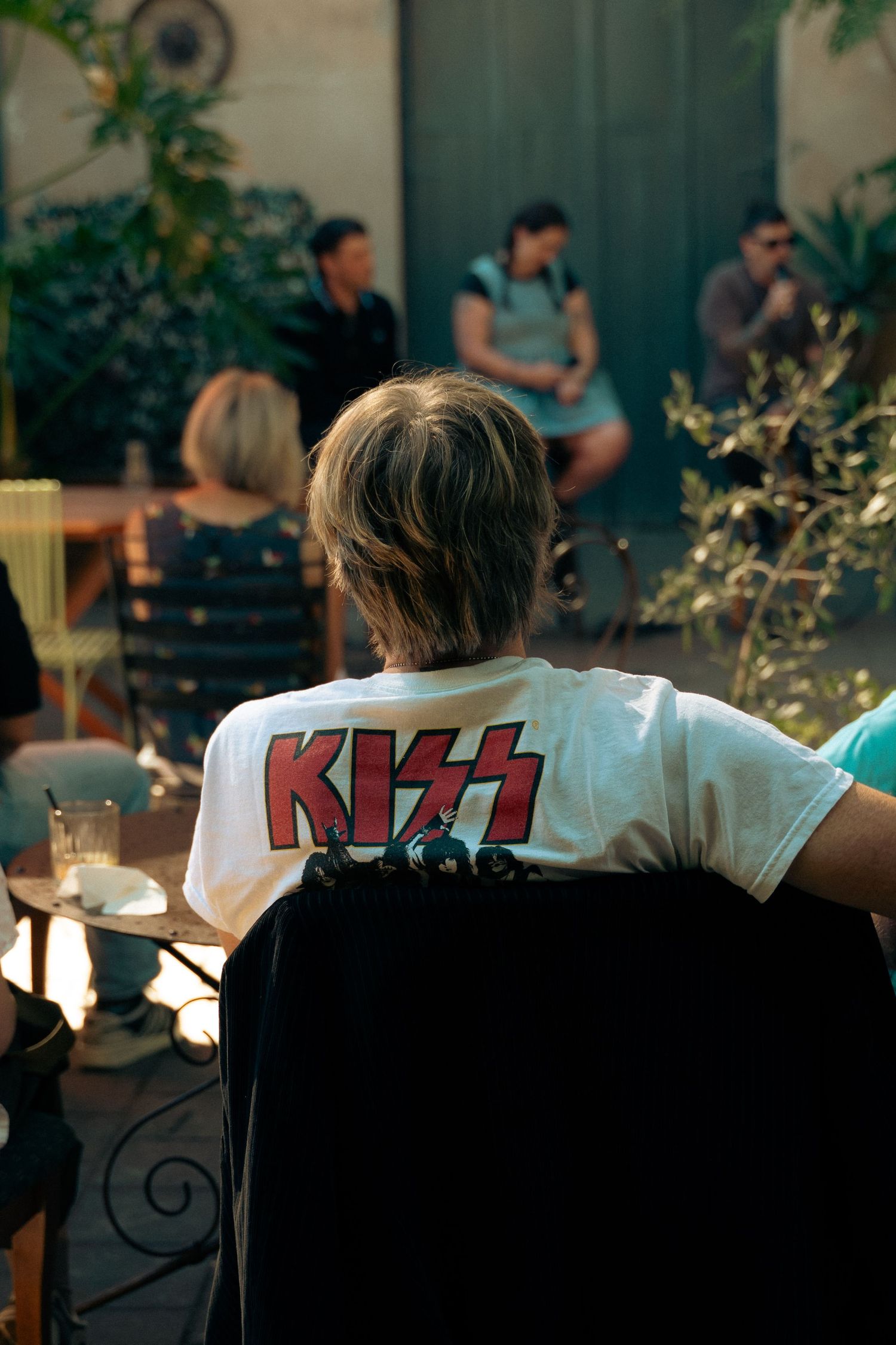 Person wearing white KISS band t-shirt seated at outdoor gathering with people in background.