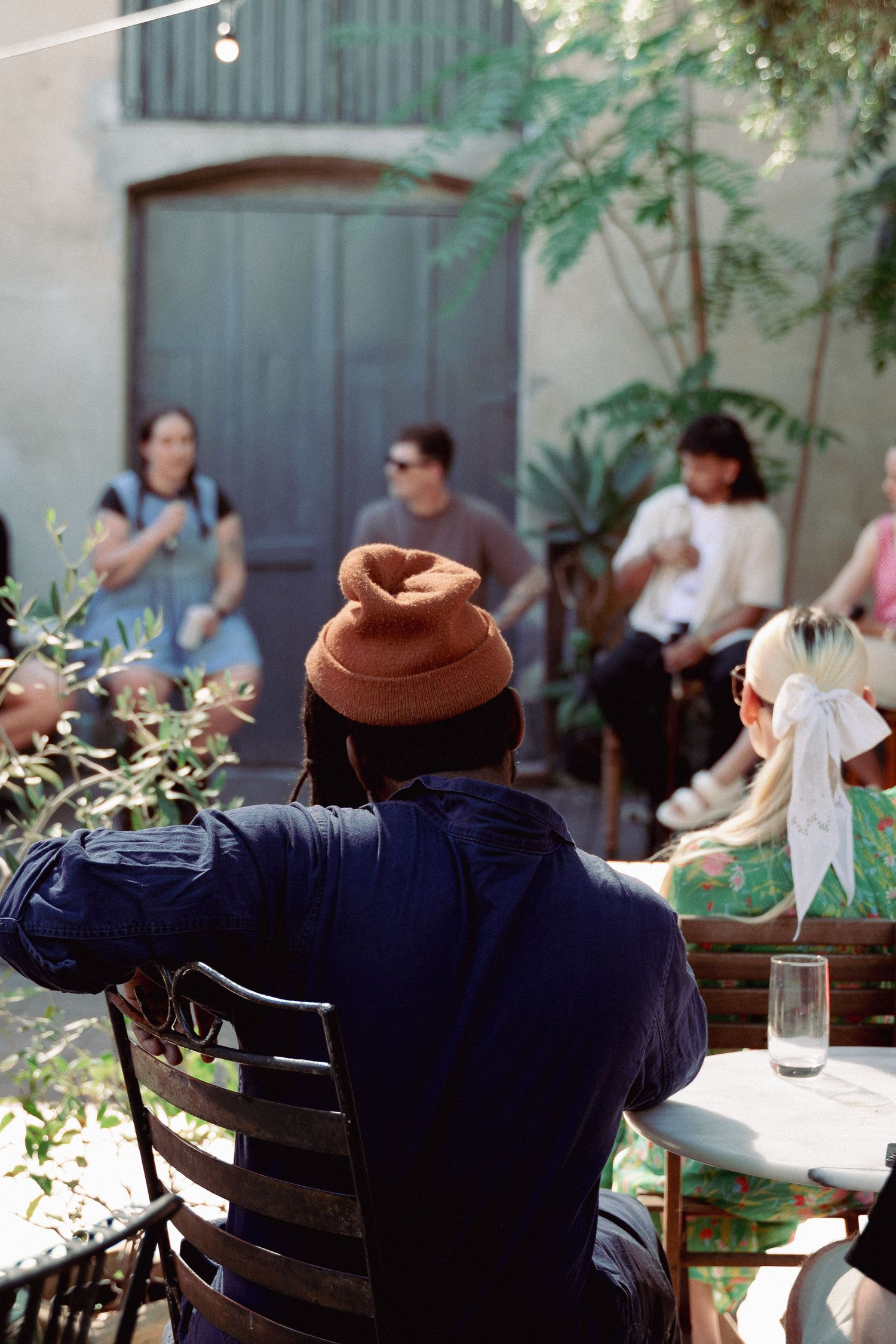 Person in orange beanie and navy jacket seated at outdoor cafe with group of people in background.