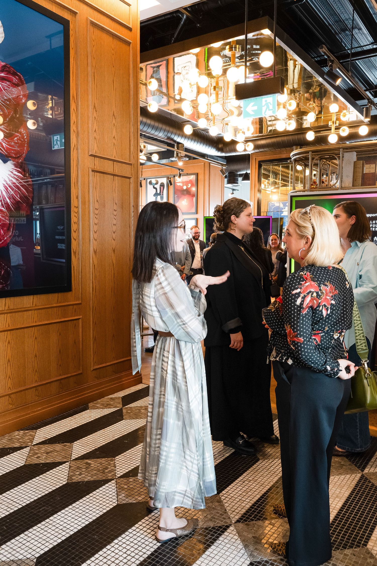 Women networking in a stylish venue with warm lighting, wood paneling, and black-and-white tiled floors.
