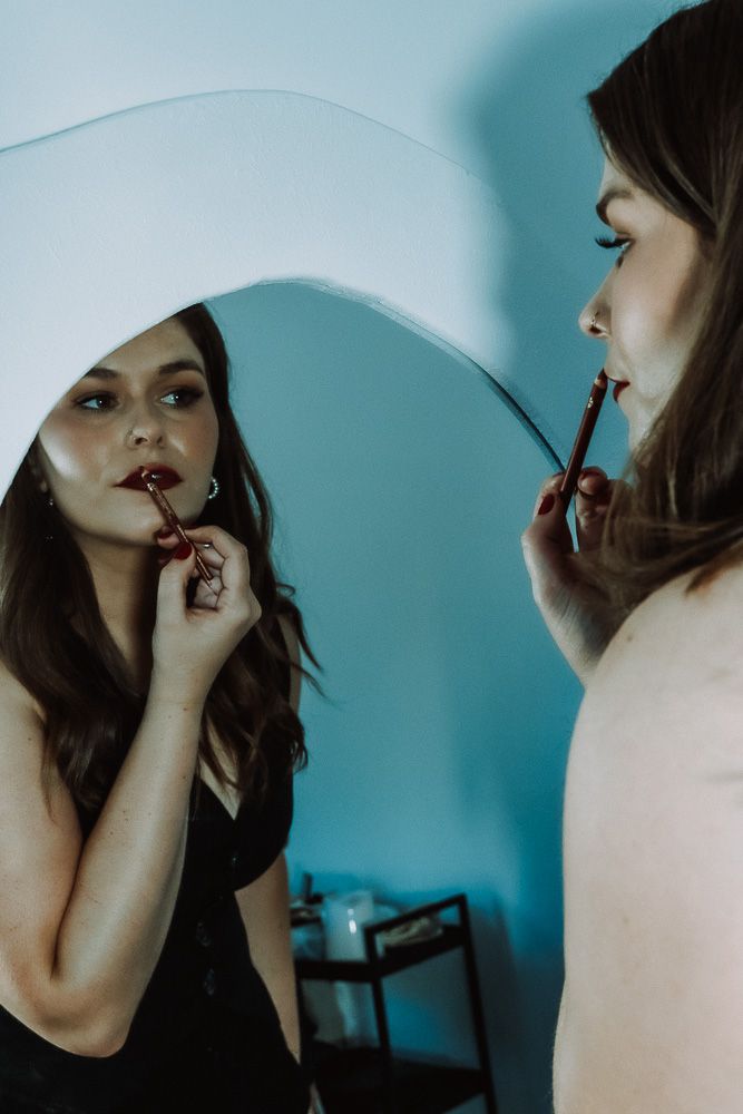 Woman applying dark red lipstick while looking in a round mirror against a blue wall.