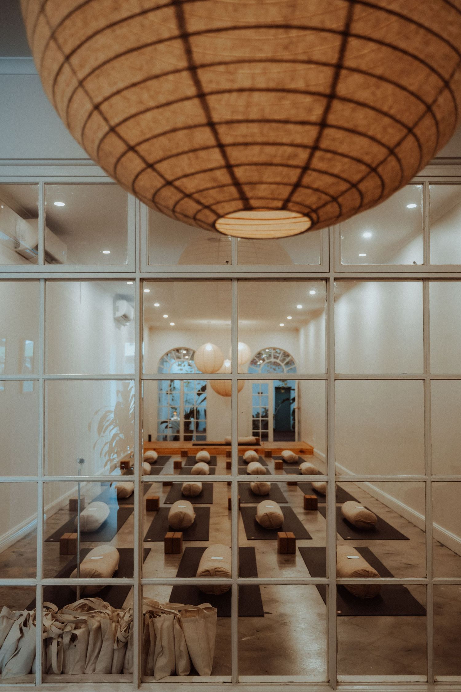 A serene yoga studio with neatly arranged mats and bolsters, viewed through glass walls with a large woven pendant light overhead.