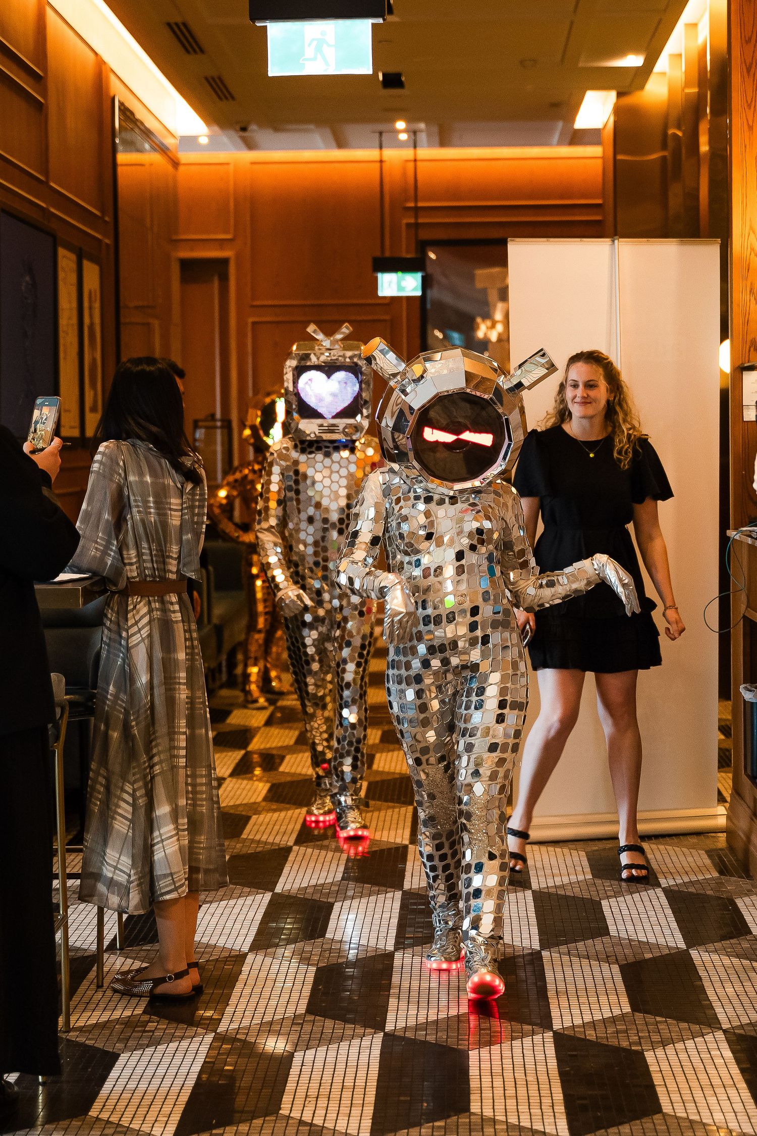 Two performers in mirrored disco ball costumes walk through an elegant hotel corridor at a glamorous event.