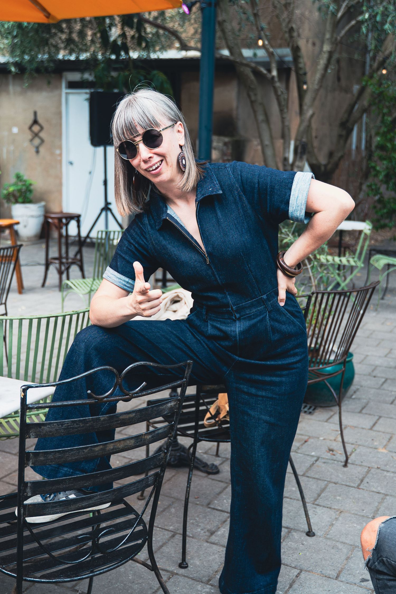 Woman in denim jumpsuit posing outdoors at patio restaurant, wearing sunglasses and giving thumbs up.