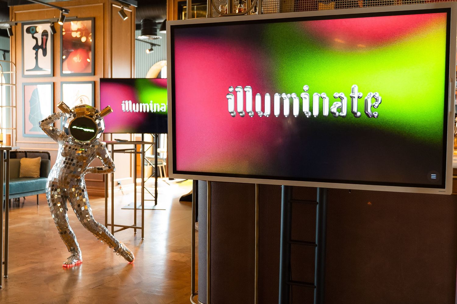 A performer in a silver sequin suit dances near illuminated screens displaying the word Illuminate at an indoor event.